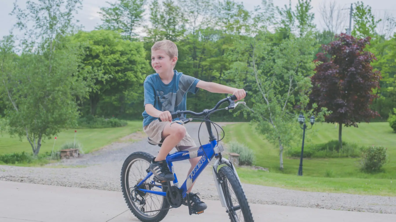 Un niño pequeño conduce una bicicleta azul por un camino pavimentado, rodeado de árboles y césped frondosos en un día despejado. Lleva una camiseta azul y pantalones cortos beige y sonríe mientras pedalea hacia adelante.