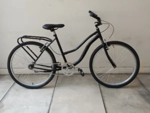 Una bicicleta PLAYERA NEGRA está estacionada sobre un piso de baldosas de color claro contra una pared blanca lisa, presenta un acabado negro elegante con un cuadro de acceso bajo, portaequipaje trasero y dos ruedas grandes. También cuenta con un asiento cómodo y empuñaduras de goma en el manillar para una mejor experiencia de conducción.
