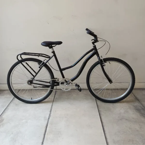 Una bicicleta PLAYERA NEGRA está estacionada sobre un piso de baldosas de color claro contra una pared blanca lisa, presenta un acabado negro elegante con un cuadro de acceso bajo, portaequipaje trasero y dos ruedas grandes. También cuenta con un asiento cómodo y empuñaduras de goma en el manillar para una mejor experiencia de conducción.