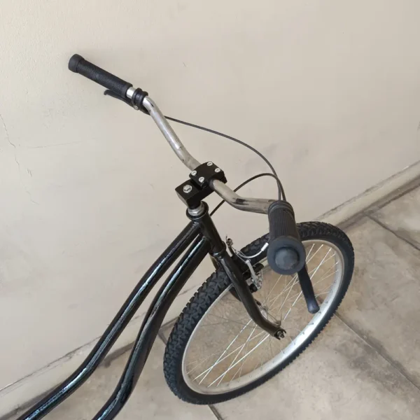 Una bicicleta PLAYERA NEGRA con un manillar curvado y puños desgastados está colocada sobre un suelo de baldosas contra una pared de color beige. La foto se centra en la mitad delantera de la bicicleta, mostrando la rueda delantera y la palanca del freno.