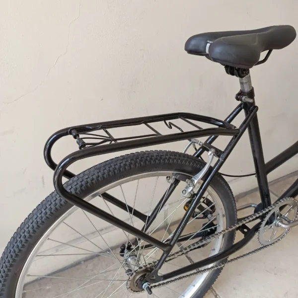 Primer plano de la parte trasera de la bicicleta PLAYERA NEGRA, que muestra un resistente portaequipaje de metal sobre la rueda trasera. La bicicleta cuenta con un cómodo asiento negro y neumáticos texturizados, y está apoyada contra una pared de color claro.